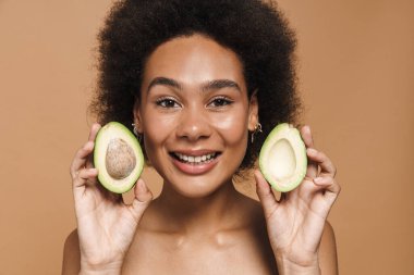 Siyah kıvırcık tişörtsüz kadın, bej arka planda izole edilmiş avokadoyla poz verirken gülümsüyor.