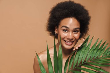 Bej arka planda yeşil yapraklı poz verirken gülümseyen siyah tişörtsüz kadın