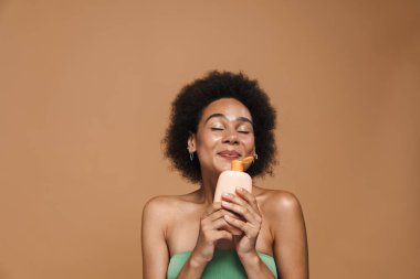 Siyah esmer kadın, bej arka planda izole edilmiş güzellik ürünüyle poz verirken gülümsüyor.