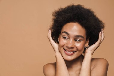 Siyah kıvırcık tişörtsüz kadın gülümsüyor ve saçlarını bej arka planda tek başına tutuyor.