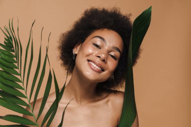 Black shirtless woman smiling while posing with green leaves isolated over beige background