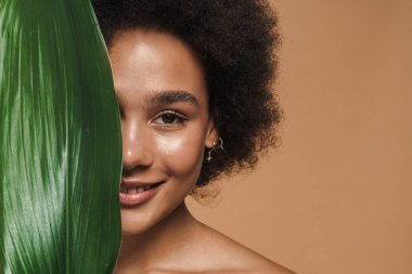 Bej arka planda yeşil yapraklı poz verirken gülümseyen siyah tişörtsüz kadın