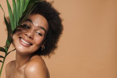 Bej arka planda yeşil yapraklı poz verirken gülümseyen siyah tişörtsüz kadın