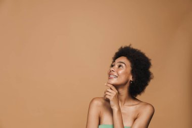 Siyah esmer kıvırcık kadın gülümsüyor ve bej arka planda tek başına yukarıya bakıyor.