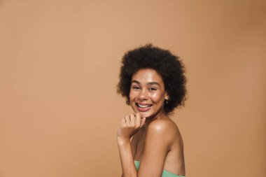 Siyah esmer kıvırcık kadın gülümsüyor ve bej arka planda izole edilmiş kameraya bakıyor.