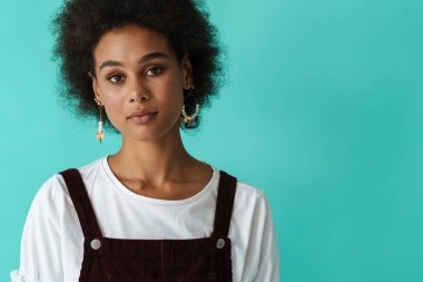 Black young woman in earrings posing and looking at camera isolated over blue background