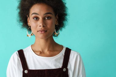 Küpeli siyahi bir kadın mavi arka planda poz veriyor ve kameraya bakıyor.