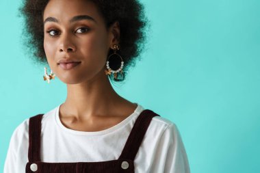 Küpeli siyahi bir kadın mavi arka planda poz veriyor ve kameraya bakıyor.