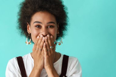 Küpe takan genç siyahi bir kadın mavi arka planda, ağzını kapatıyor.