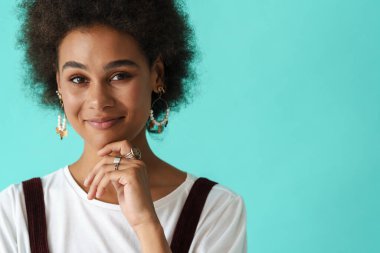 Küpe takan genç siyah kadın gülümsüyor ve mavi arka planda izole edilmiş kameraya bakıyor.