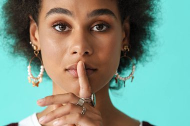 Küpeli siyahi genç bir kadın, mavi arka planda izole edilmiş kameraya sessiz jest yapıyor.