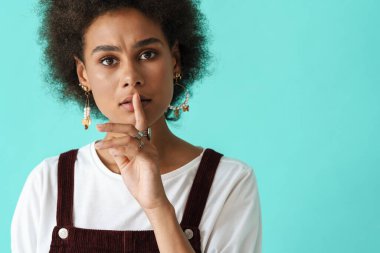 Küpeli siyahi genç bir kadın, mavi arka planda izole edilmiş kameraya sessiz jest yapıyor.