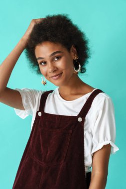 Küpeli siyahi bir kadın gülümsüyor ve mavi arka planda izole edilmiş kameraya bakıyor.