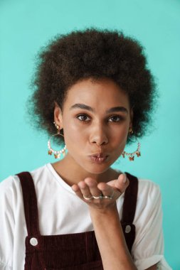 Küpe takan genç siyahi kadın mavi arka planda izole edilmiş kameraya hava öpücüğü veriyor.