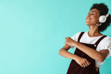 Mavi arka planda izole edilmiş kulaklıklarla müzik dinlerken dans eden genç zenci kadın.