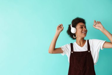 Mavi arka planda izole edilmiş kulaklıklarla müzik dinlerken dans eden genç siyah bir kadın.
