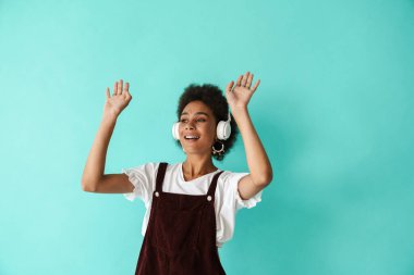 Mavi arka planda izole edilmiş kulaklıklarla müzik dinlerken dans eden genç siyah bir kadın.