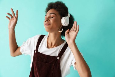 Mavi arka planda izole edilmiş kulaklıklarla müzik dinlerken dans eden genç siyah bir kadın.