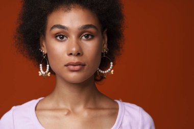Black young woman in earrings posing and looking at camera isolated over brown background