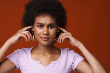 Young black woman wearing earrings frowning while plugging her ears isolated over brown background
