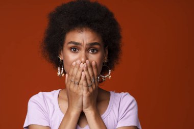 Young black woman frowning while covering her mouth isolated over brown background