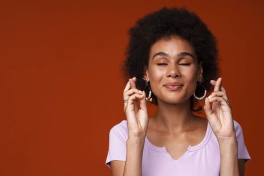 Genç siyah kadın gülümserken parmaklarını çapraz yapıp kahverengi arka planda izole bir şekilde poz veriyor.