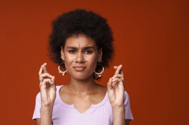 Young black woman frowning while posing with fingers crossed isolated over brown background