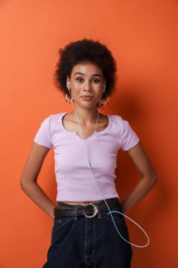 Küpeli genç bir kadın kahverengi arka planda izole edilmiş kulaklıklı müzik dinliyor.