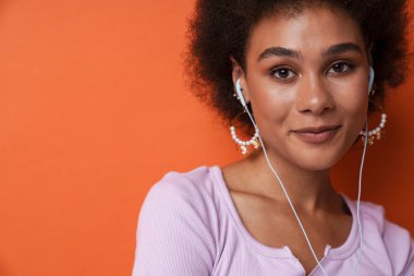 Genç siyahi kadın turuncu arka planda izole edilmiş kulaklıkla müzik dinlerken gülümsüyor.