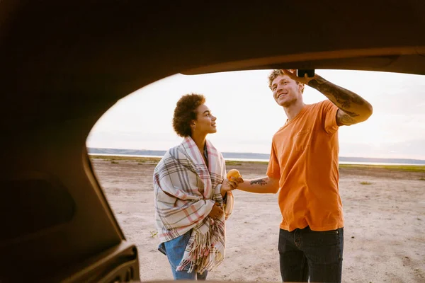 Araba gezisi sırasında bagajda durup gülümseyen ve konuşan çok kültürlü genç bir çift.