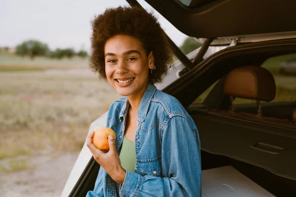 Young multiracial woman eating peach while sitting in trunk during car trip