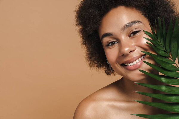 Black shirtless woman smiling while posing with green leaf isolated over beige background