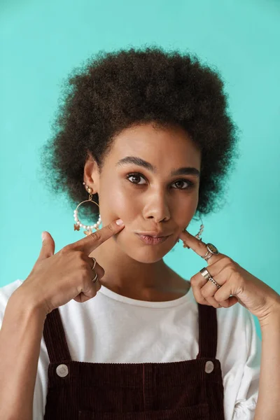 Genç siyah kadın gülümsüyor ve parmaklarını yanaklarını işaret ediyor mavi arka planda soyutlanmış.