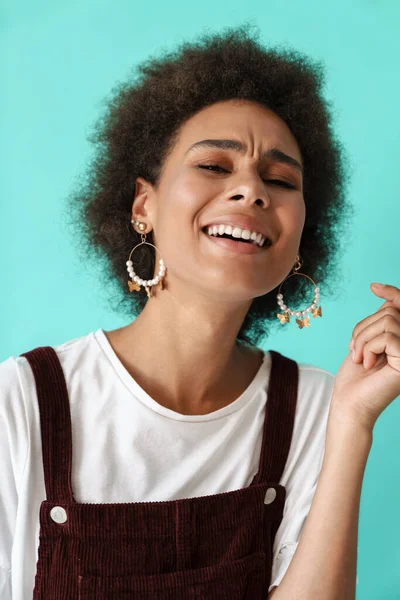 Küpe takan genç siyah kadın gülüyor ve mavi arka planda izole edilmiş kameraya bakıyor.