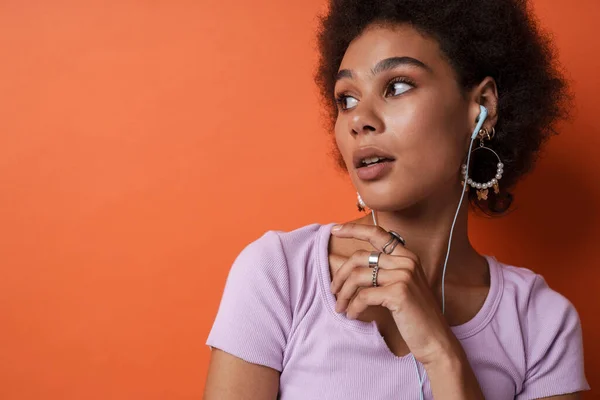 Young black woman wearing earrings listening music with earphones isolated over orange background