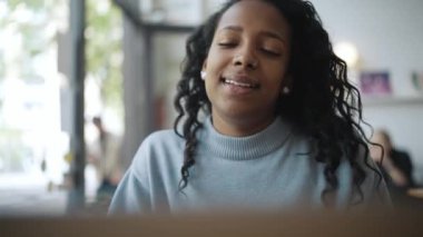 Happy African woman wearing blue sweater talking by video call on laptop in cafe