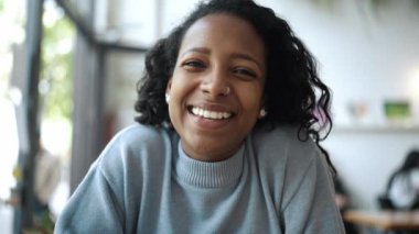 Mavi kazak giyen gülümseyen Afrikalı kadın laptopla konuşuyor ve kafede karşılama jesti yapıyor.