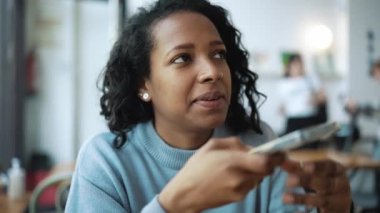 Mavi kazak giyen gülümseyen Afrikalı kadın kafede telefonla sesli mesaj kaydediyor.