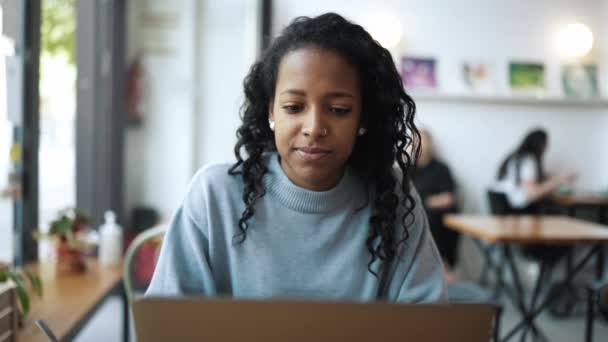 穿着蓝色毛衣在咖啡店用笔记本电脑打字的多愁善感的非洲女人 — 图库视频影像