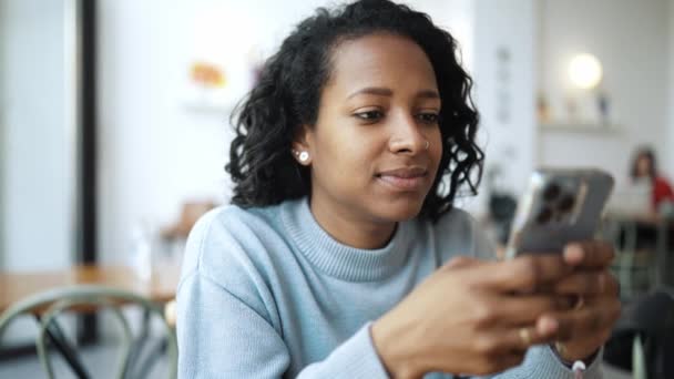 非洲女人穿着蓝色毛衣 在电话里打字 在咖啡店里看着相机 — 图库视频影像
