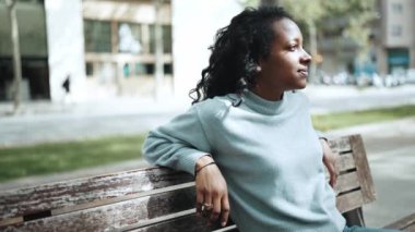 Mavi kazak giyen, açık hava bankında oturan, mutlu, esmer Afrikalı kadın.