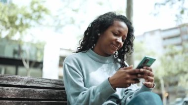 Mavi kazak giyen mutlu, esmer bir kadın açık hava bankında cep telefonuyla yazı yazıyor.