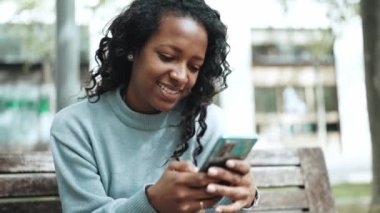 Neşeli esmer Afrikalı kadın mavi kazak giyiyor açık hava bankında cep telefonuyla yazı yazıyor.