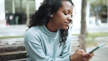 Gülümseyen esmer Afrikalı kadın mavi kazak giyiyor açık hava bankında cep telefonuyla yazı yazıyor.