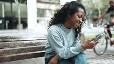 Kahkahalar atan esmer Afrikalı kadın mavi kazak giyiyor açık hava bankında cep telefonuyla yazı yazıyor.