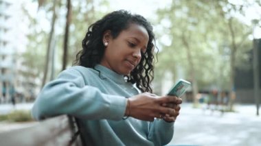 Güzel esmer Afrikalı kadın mavi süveter giyiyor açık hava bankında cep telefonuyla yazı yazıyor.