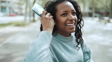 Kahkahalar atan esmer Afrikalı kadın mavi kazak giyiyor. Dışarıdaki bankta cep telefonundan sesli mesaj kaydediyor.