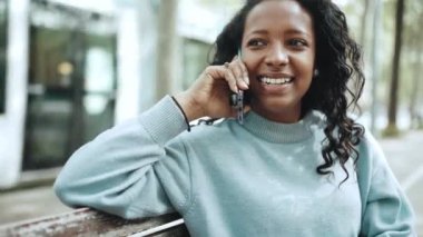 Mavi kazak giyen kendine güvenen esmer Afrikalı kadın açık hava bankında cep telefonuyla konuşuyor.