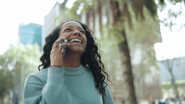 Kahkaha atan esmer Afrikalı kadın mavi kazak giyiyor telefonla konuşuyor ve sokakta yürüyor.