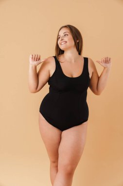 Mayo giyen, gülümseyen ve kendini işaret eden genç bir kadın bej duvarın üzerinden izole edilmiş.